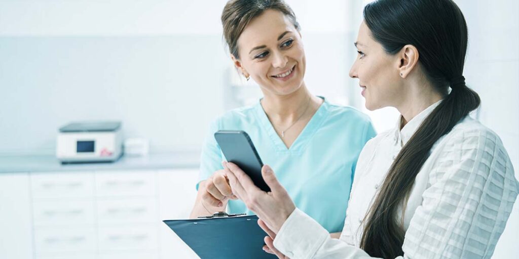 Mulher mostrando o celular para médica sorridente em seu consultório- E-clínica marketing digital